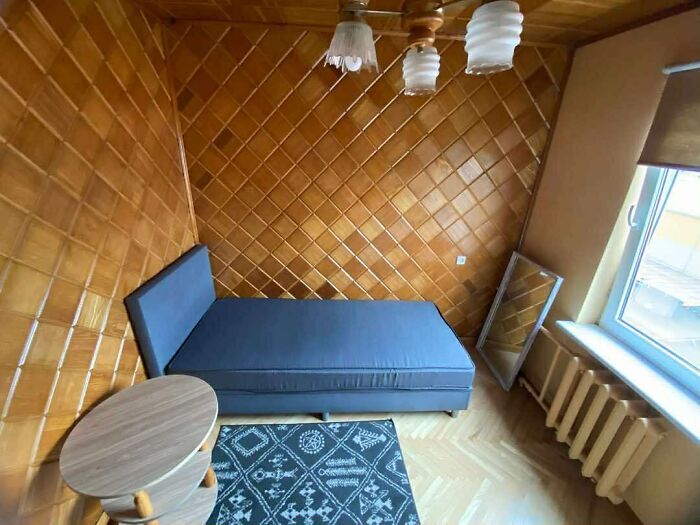 Small bedroom with dark blue bed, wooden side table, black patterned rug, and angled wood-paneled walls in real estate listing.