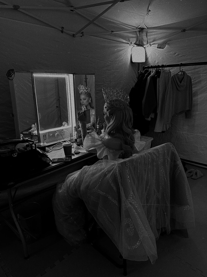 Ariana Grande in a backstage dressing room wearing a detailed gown and crown, reflecting on emotional age regression.