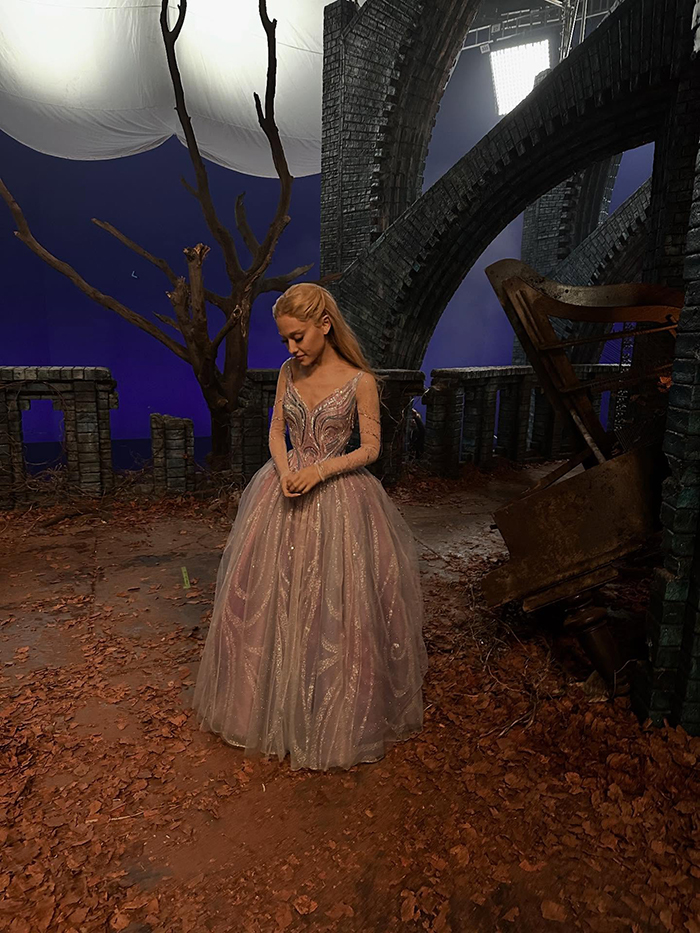 Ariana Grande wearing a sparkling gown on a film set with autumn leaves and stone arches, hinting at emotional age regression.
