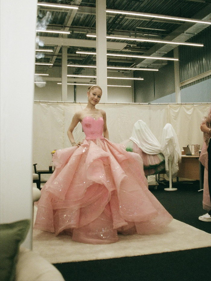 Ariana Grande wearing a voluminous pink gown in a backstage room, highlighting emotional age regression theme.
