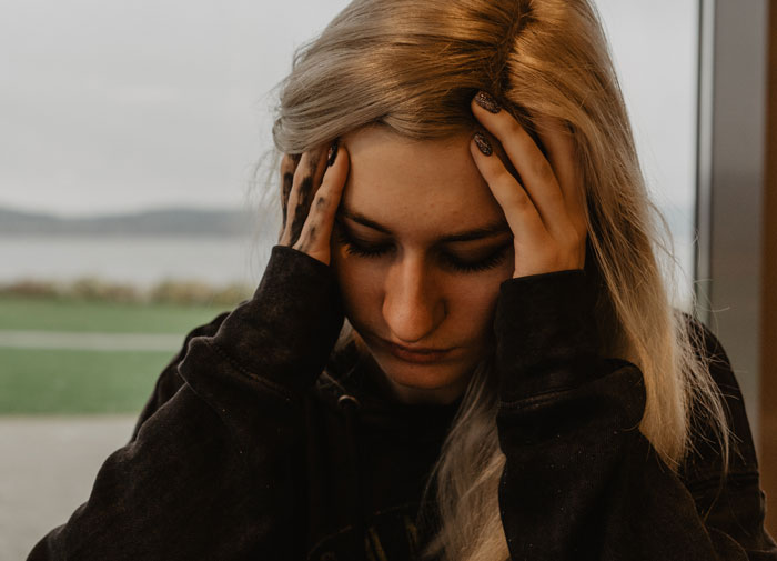 A woman with long blonde hair holding her head, looking upset indoors near a window with an outdoor view.