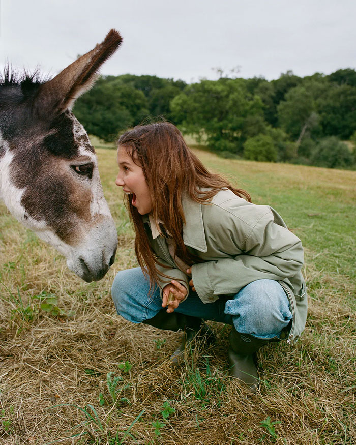 Millie Bobby Brown in a green coat crouching outdoors, engaging closely with a donkey in a rural grassy field. Millie Bobby Brown in a green coat crouching outdoors, engaging closely with a donkey in a rural grassy field.