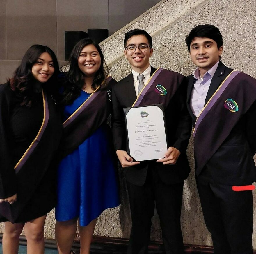 Four young Asian graduates smiling, dressed in formal attire with AIM sashes, celebrating academic achievement together.