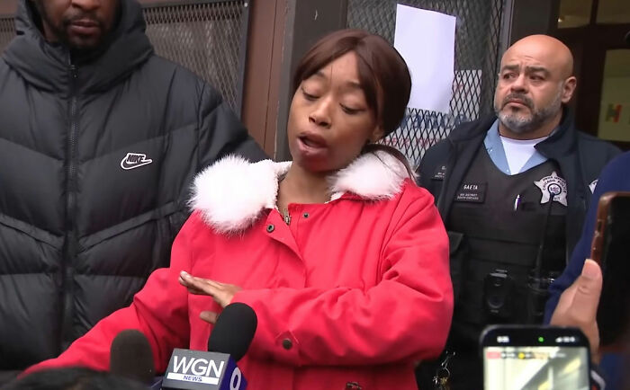 Woman in red coat speaking to media after pregnant woman is attacked by young school kids, with police present nearby.