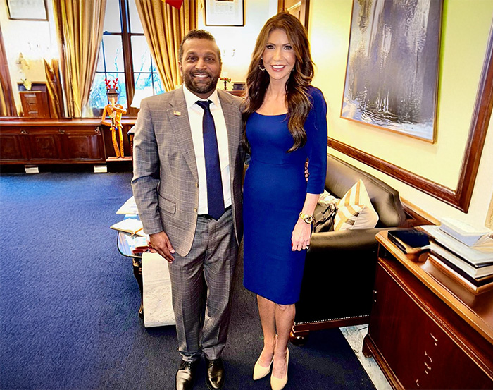 Kristi Noem standing in a blue dress beside a man in a suit in an office, showcasing her transformation over the years. Kristi Noem standing in a blue dress beside a man in a suit in an office, showcasing her transformation over the years.