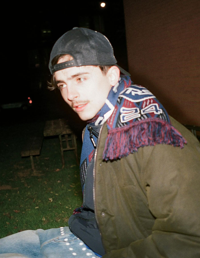 Young man wearing a backward cap and a patterned scarf sitting outdoors at night, linked to Timoth&eacute;e Chalamet and EsDeeKid theory.