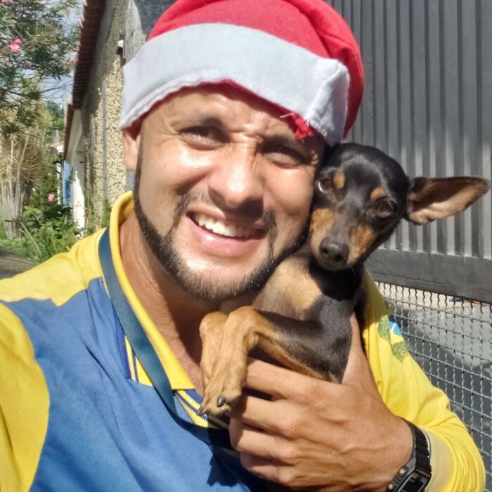 Mailman wearing a red hat smiling and holding a small dog while standing outdoors near a fence on his route.