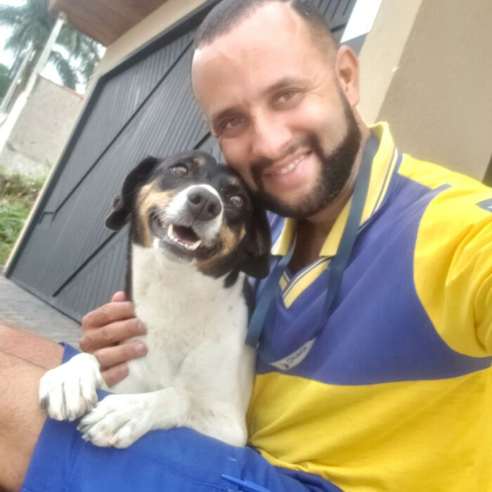 Mailman wearing yellow and blue uniform smiling and sitting with a happy black and white dog on his route outdoors.