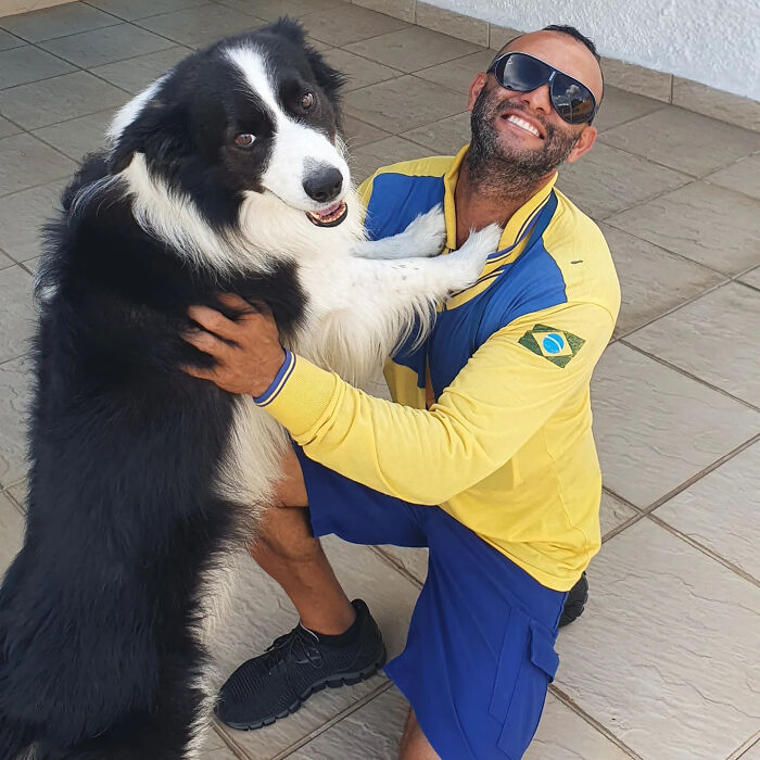 Mailman wearing yellow and blue uniform happily hugging a friendly black and white dog on his route.