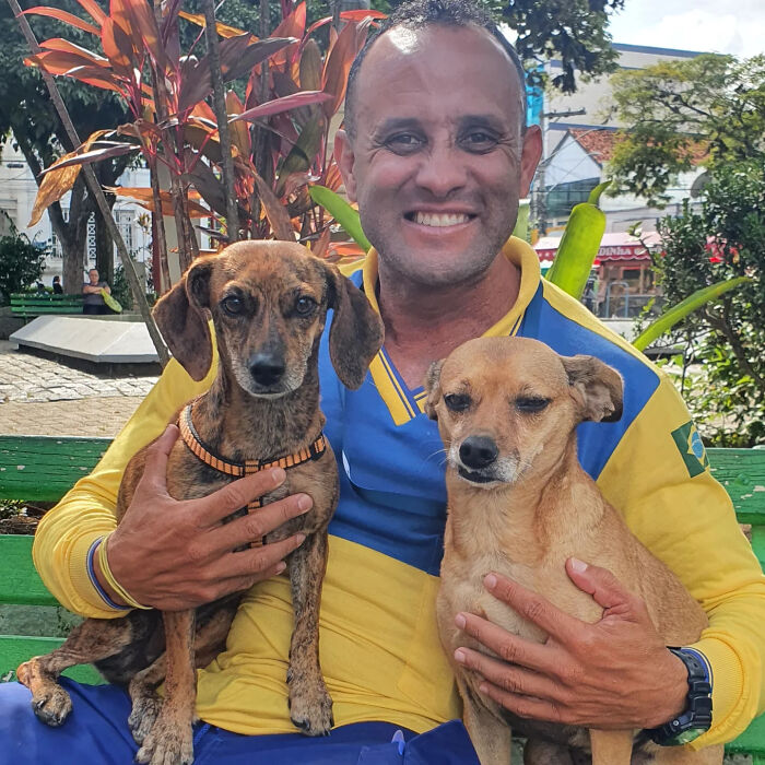 Mailman sitting on a bench outdoors, smiling and holding two dogs, showing his bond with dogs on his route.