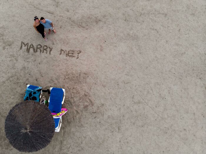 Couple on the beach smiling next to a marriage proposal written in the sand, showcasing faith in humanity restored.
