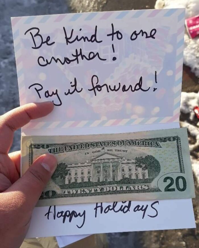 Hand holding a holiday card with a 20-dollar bill and a note encouraging kindness and paying it forward.