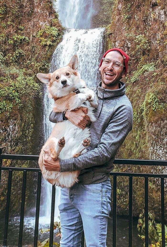 Man holding a corgi smiling in front of a waterfall, capturing a moment of faith in humanity restored.