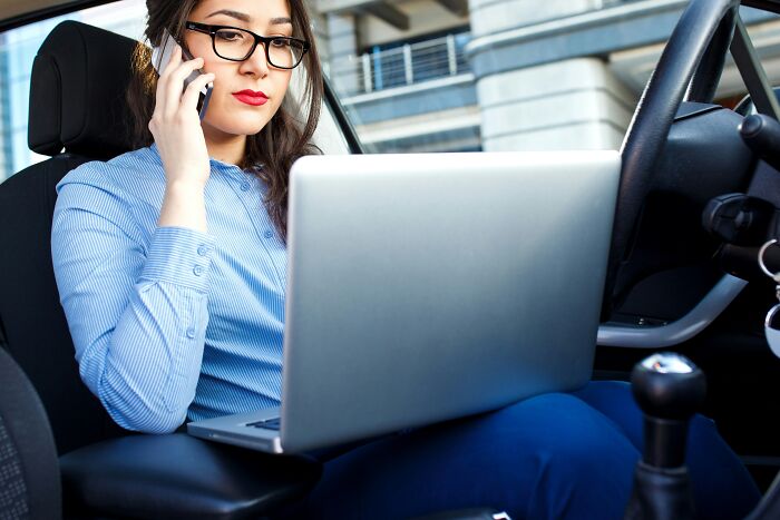 Woman in blue shirt sitting in car using laptop and phone, focused on identifying toxic relationship traits.
