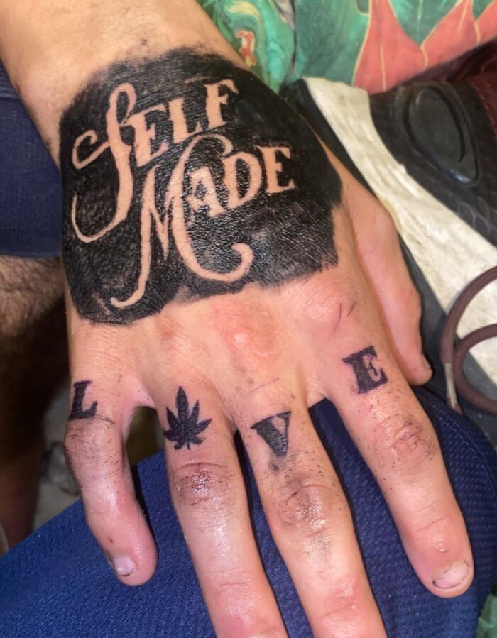 Hand tattoo with large black ink reading self made and fingers tattooed with letters and a leaf symbol seen up close.
