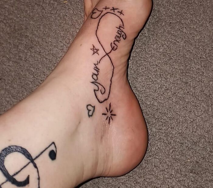 Foot tattoo featuring an infinity symbol with the words your enough and small decorative stars and hearts inked on the skin.