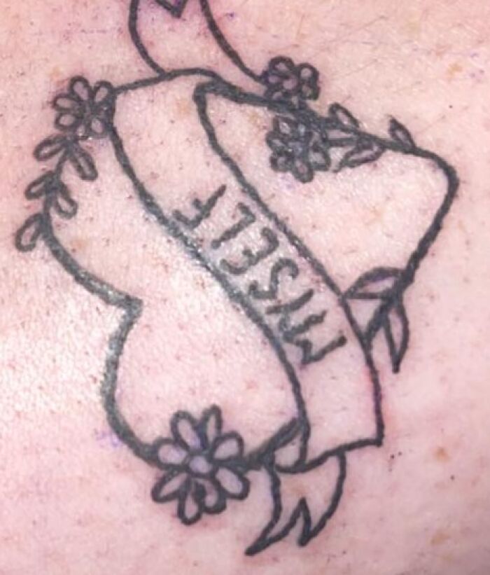 Simple black ink tattoo of a broken heart with flowers and a misspelled banner on light skin in unusual tattoo designs.