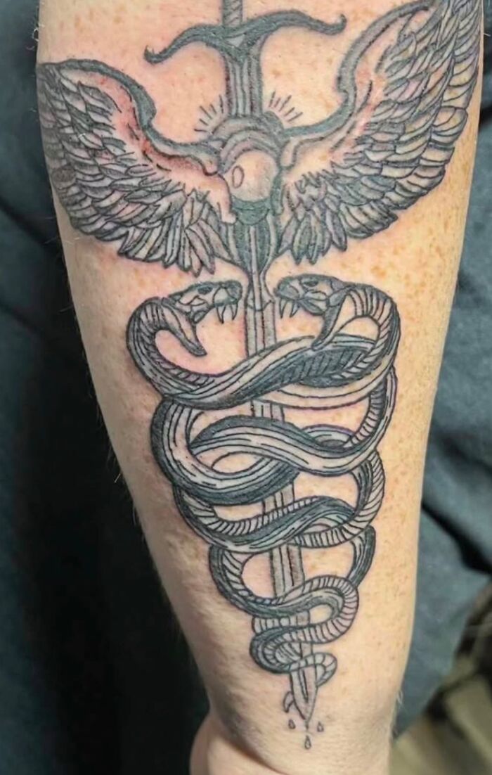 Tattoo of a sword with wings and two snakes wrapped around it on a forearm, unusual body tattoo design.