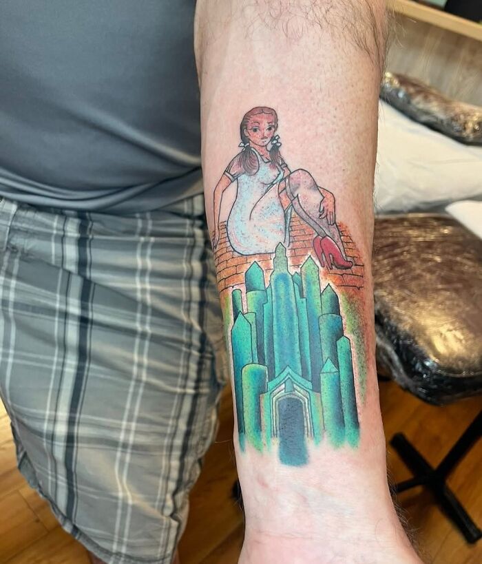 Forearm tattoo of a woman sitting on a brick wall above a colorful cityscape in a unique and bold tattoo design.