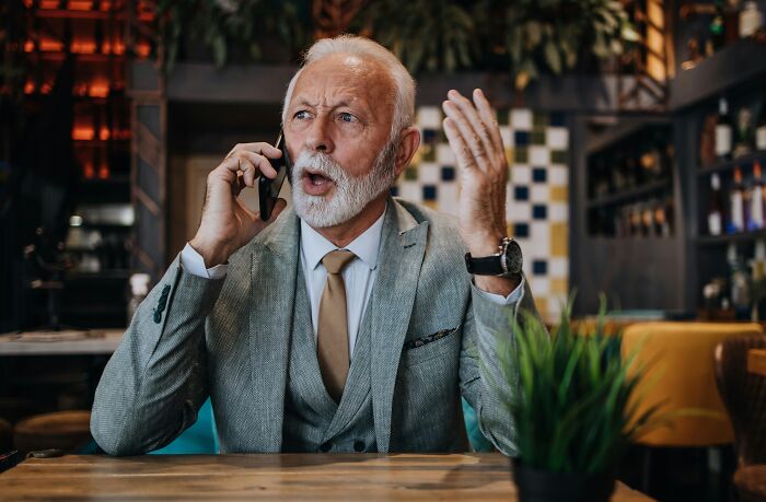 Elderly man in a gray suit talking on phone showing frustration, illustrating toxic relationship traits in a cozy cafe setting.