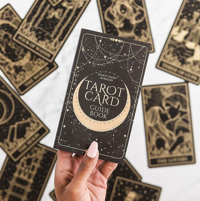 Hand holding a tarot card guide book with tarot cards spread out, a popular hobby to keep hands busy during hibernation season.