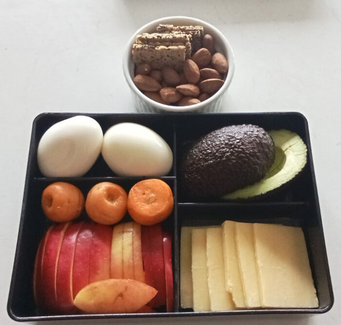 Girl dinner combo with boiled eggs, avocado, cheese slices, apple slices, carrots, almonds, and crackers in a black container.