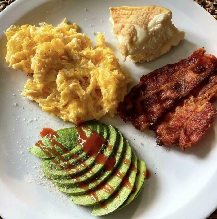 Plate with girl dinner combos including scrambled eggs, sliced avocado with sauce, bacon, and biscuit with butter.