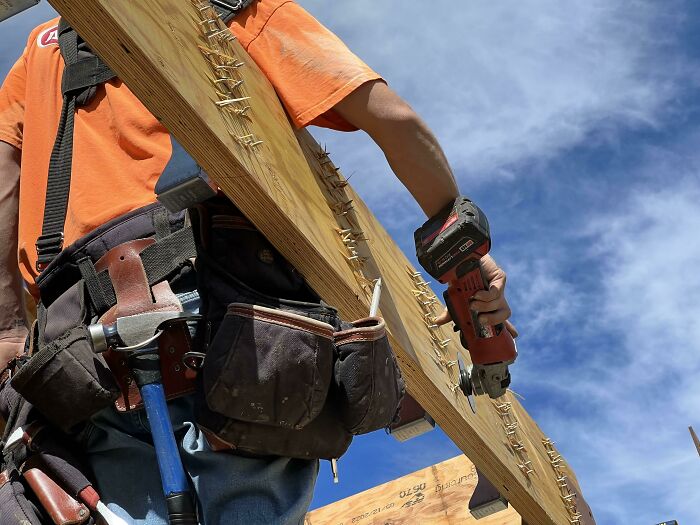 Construction worker carrying a wooden plank with protruding nails, showcasing zero common sense safety risk outdoors.