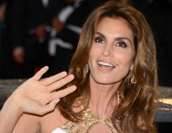 Style icon with flowing brown hair, wearing a gold-embellished dress, smiling and waving at an event.