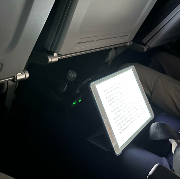 Tablet glowing brightly under airplane tray table in dark cabin, illustrating people with zero common sense on flights.