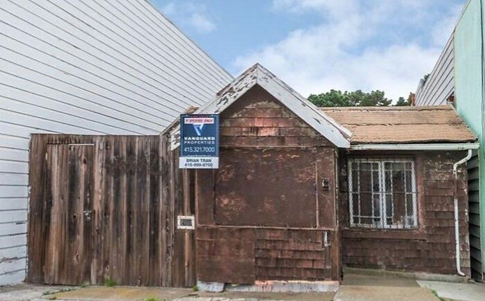 Small weathered Zillow home with a for sale sign between two larger buildings under a blue sky.