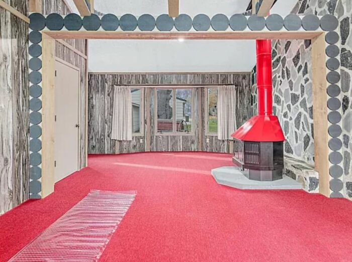 Unique Zillow homes with bold red carpet and retro fireplace in a wood-paneled living room with large windows.