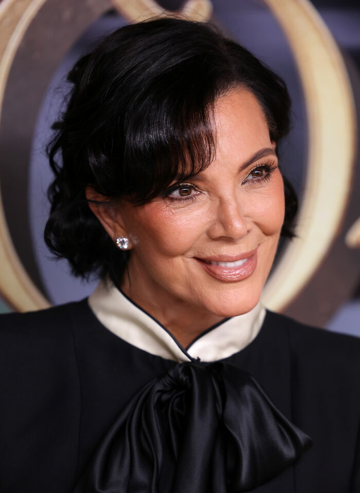 Kris Jenner smiling at an event, wearing a black outfit with a satin bow and diamond stud earrings.