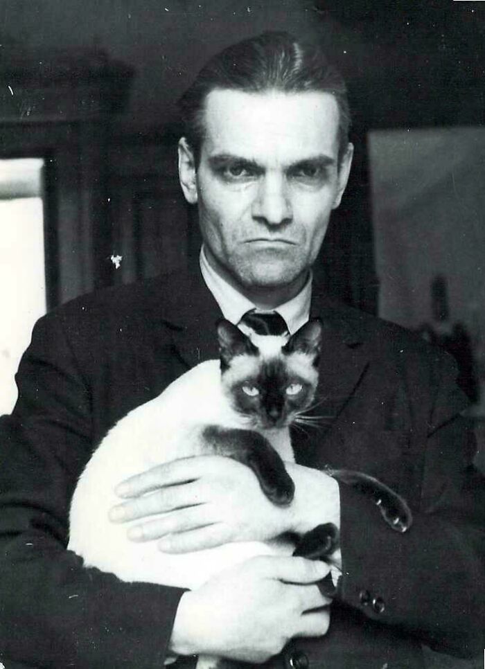 Black and white unique historical photo of a stern man holding a Siamese cat indoors with a serious expression.