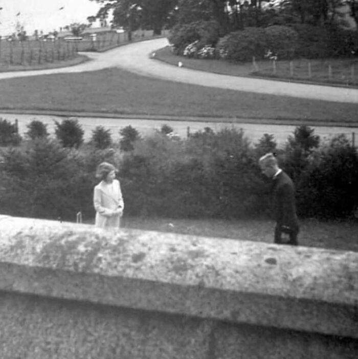 Black and white unique historical photo showing two people in formal attire outdoors near bushes and a curved road.