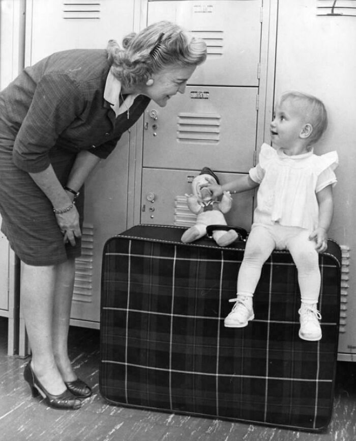 Black and white historical photo of a woman smiling at a toddler sitting on a large plaid suitcase with lockers behind.