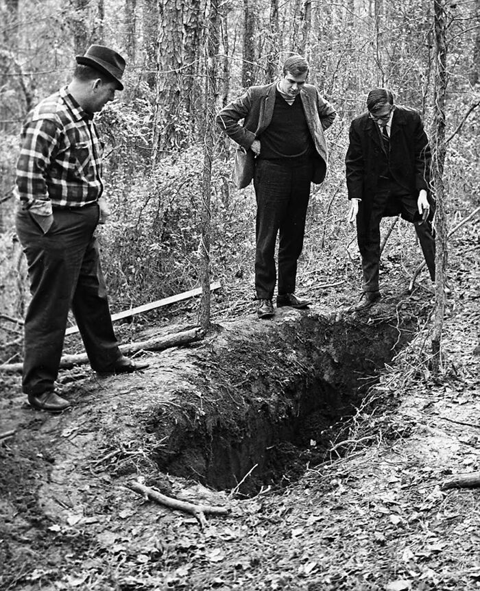 Three men in vintage clothing standing around a freshly dug hole in the forest in a unique historical photo.