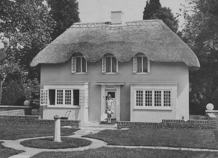 Black and white photo of a woman standing in front of a unique historical thatched-roof house with a garden path.