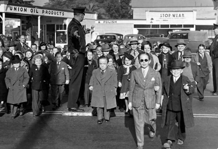 Group of children dressed in vintage suits crossing street with police officer directing traffic in a unique historical photo.