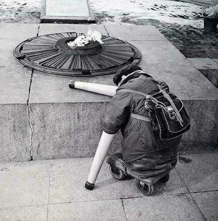 Black and white historical photo of a child with prosthetic legs kneeling by a memorial flame, showing unique resilience.