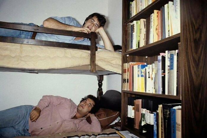 Two men resting on bunk beds in a room with a bookshelf filled with books, a unique historical photo with deep backstory.