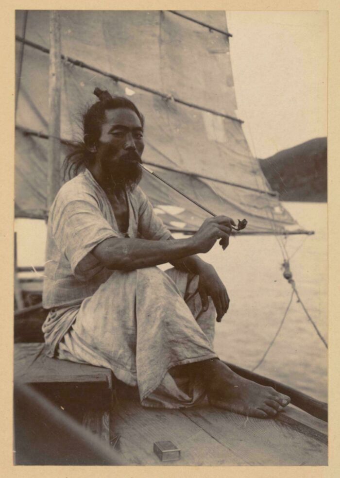 Man in traditional clothing smoking a pipe while sitting on a boat, a unique historical photo with deep backstory.