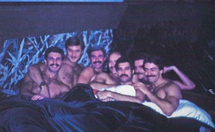Group of shirtless men with mustaches lying closely together under blankets in a unique historical photo.