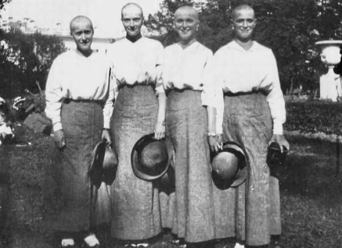 Four smiling women with shaved heads, dressed in long skirts and holding hats, in a historical outdoor photo.