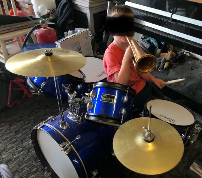 Child playing a blue drum set while blowing a brass horn, illustrating petty revenge humor in a cluttered room.