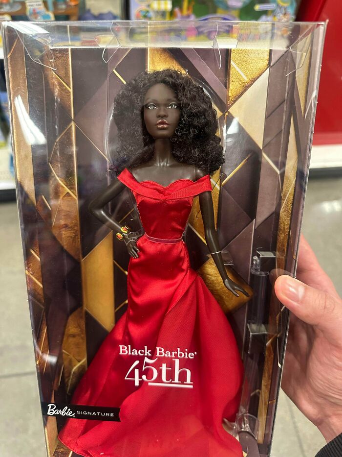 Black Barbie doll in a red dress inside packaging, held by a hand in a retail store on Black Friday.