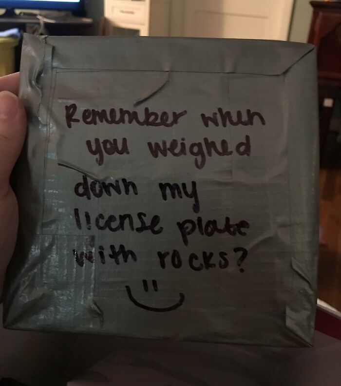 Hand holding a taped package with a handwritten note referencing petty revenge involving a license plate weighed down with rocks.