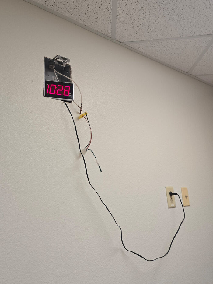 Digital clock hanging loosely on wall with exposed wires and a long power cord plugged into a distant outlet, showing a not my job fail.