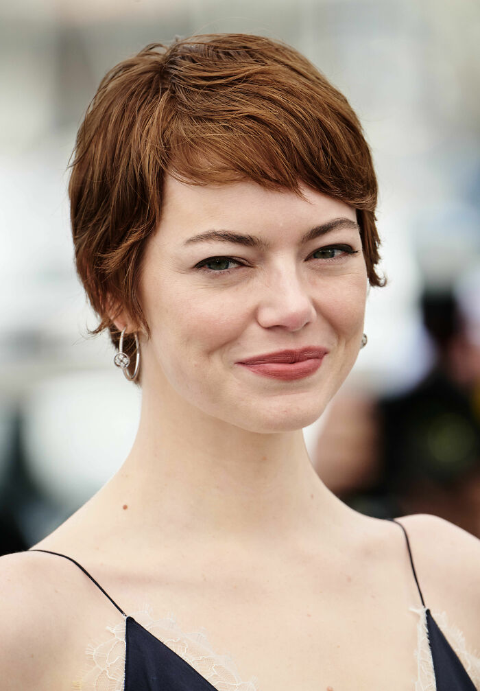 Emma Stone with short hair and earrings, smiling softly at an outdoor event, highlighting her bio and career.