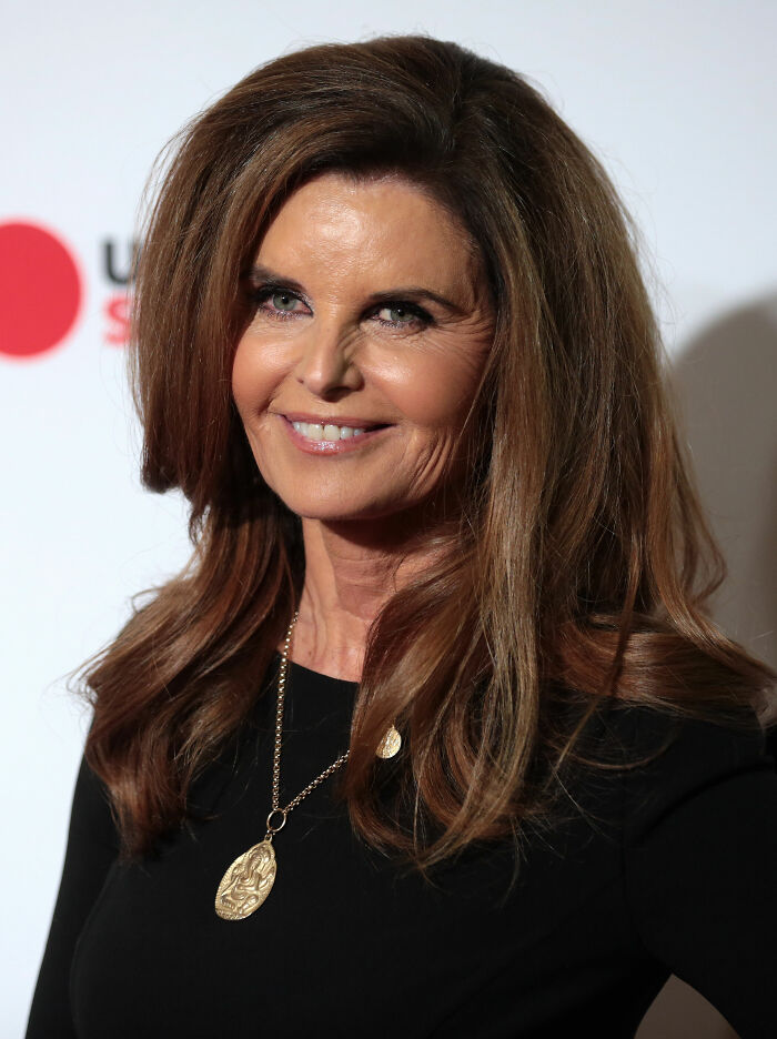 Maria Shriver smiling at an event, wearing a black top and a gold pendant necklace with styled hair.
